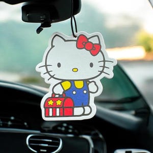 May include: A white Hello Kitty car air freshener with a red bow, yellow shirt, blue overalls, and a red and white suitcase with two stars.
