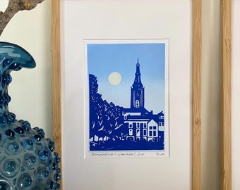 May include: A framed linocut print featuring a blue and white silhouette of a church tower and trees under a pale moon. The artwork is titled "Prinsenhof - Noordwijk, Z.H." and signed "7/26" in pencil on the white matting within a light wood frame.