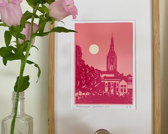 May include: A framed print featuring a pink and magenta linocut illustration of a church tower and moon, with the text "Prinseneval-Noordwijk. 8/4" and "FK 25" written at the bottom. The artwork is displayed in a light wood frame with a white mat.