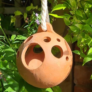 May include: A terracotta bird feeder with large circular openings and a rope for hanging. The spherical feeder is suspended among green foliage and small purple flowers, with a brick wall in the background. The feeder is designed to attract birds to a garden.
