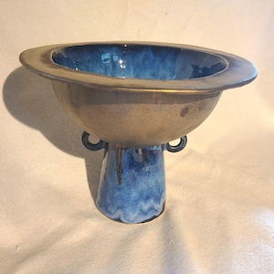 May include: A ceramic bowl with a wide, bronze-coloured rim and a deep blue interior. The bowl has a rounded body and a flared, blue base with two small handles. The glaze creates a textured, artistic effect.