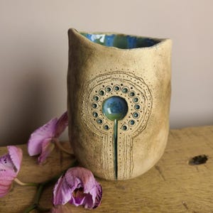 May include: A handmade ceramic vase with a textured, beige exterior. The vase features a central design with a blue and green circular element surrounded by smaller teal circles. The interior rim is blue and green. Pink orchid flowers are in the foreground.