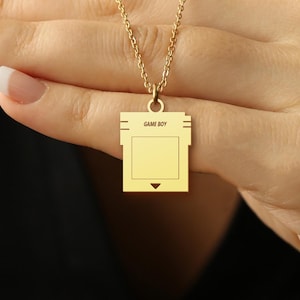 May include: A gold necklace with a Game Boy cartridge pendant. The pendant is a replica of a classic Game Boy cartridge.