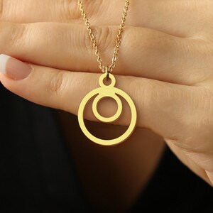 May include: A gold-colored necklace featuring a pendant with two concentric circles. The necklace is held in a hand with manicured nails against a dark background. The chain is delicate, and the pendant is smooth and shiny.