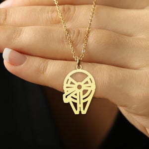 May include: A gold-colored pendant necklace featuring a stylized design. The pendant is suspended from a delicate chain. The necklace is held in a hand with manicured nails, against a dark background.