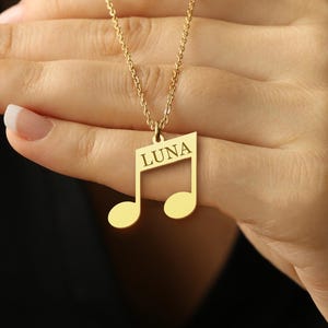 May include: A gold-colored necklace featuring a musical note pendant. The pendant has the name "LUNA" engraved on it. The necklace is held up by a hand with manicured nails, against a dark background.