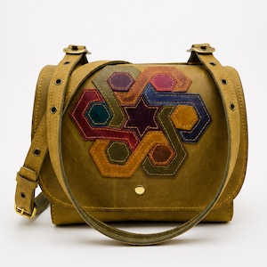 May include: An olive green leather shoulder bag featuring an adjustable strap with brass buckles and a central geometric appliqué. The decorative pattern consists of interlocking hexagonal and star shapes in shades of purple, blue, and orange.