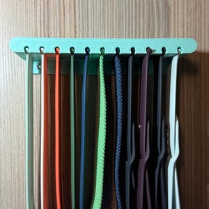 Puede incluir: Un organizador de plástico verde claro con 10 ranuras para guardar correas de reloj. El organizador está montado en una pared y tiene una variedad de correas de reloj de colores colgando.