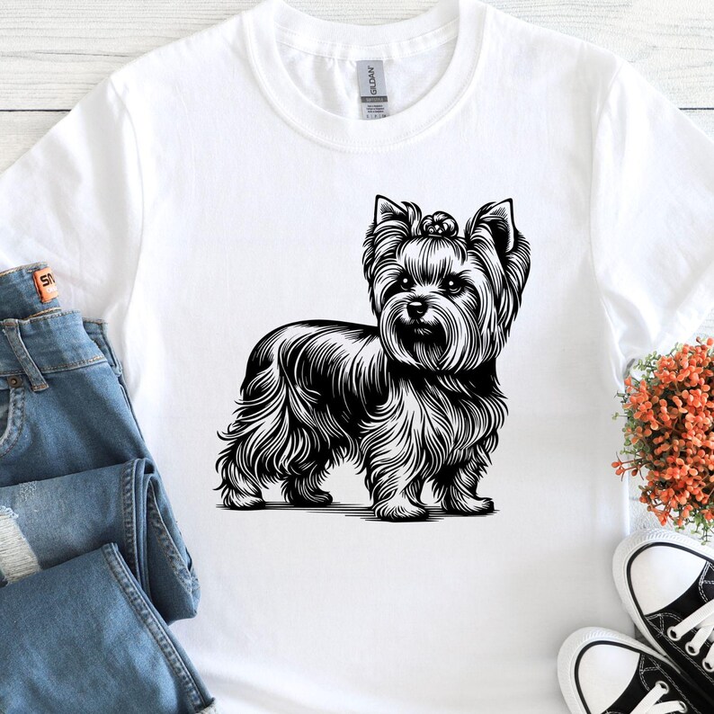 May include: White t-shirt featuring a detailed black and white illustration of a Yorkshire Terrier dog. The dog is depicted in a standing pose, showcasing its fluffy fur and distinctive features. The shirt is paired with blue jeans and black and white sneakers.