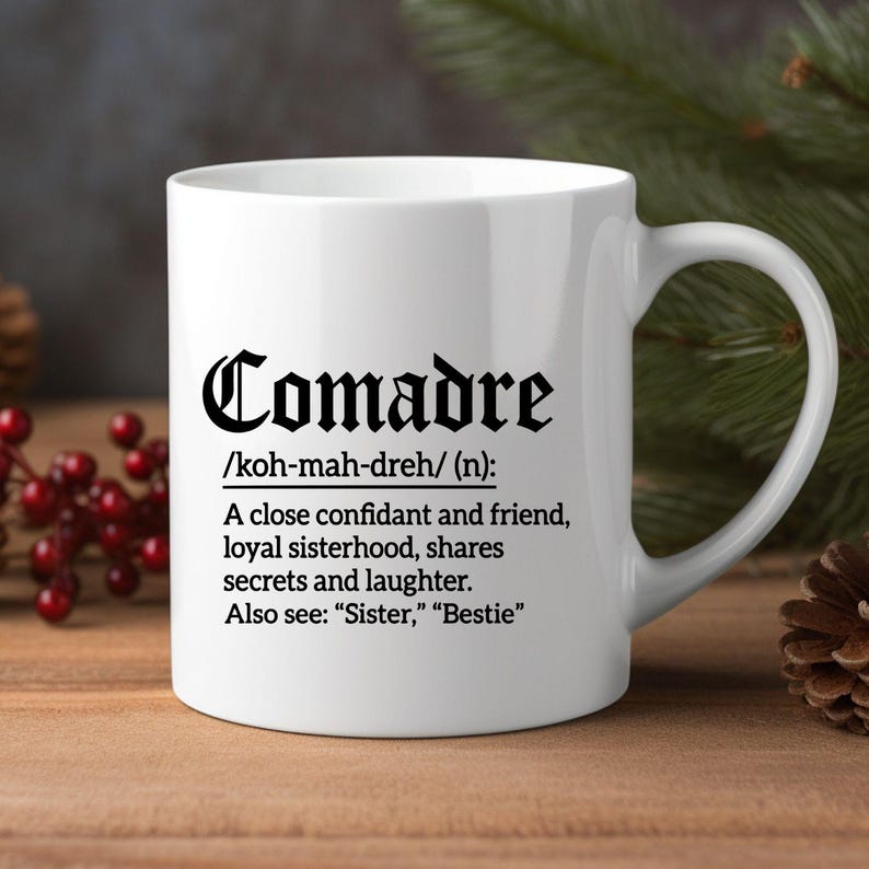 May include: White ceramic mug with the word "Comadre" in a decorative black font. Below the word is a definition: "A close confidant and friend, loyal sisterhood, shares secrets and laughter." Also see: "Sister," "Bestie".