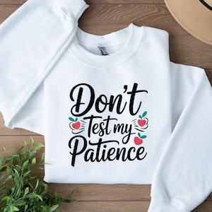 May include: White crewneck sweatshirt with the text "Don't Test my Patience" in a black script font. The design includes small red apple graphics with green leaves. The sweatshirt is folded on a wooden surface.