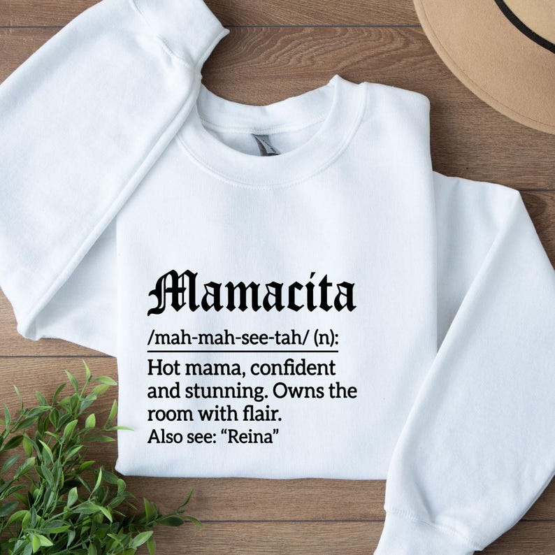 May include: White sweatshirt with the word "Mamacita" in a gothic font. Below the word is a definition: "Hot mama, confident and stunning. Owns the room with flair." The sweatshirt is on a wooden surface.