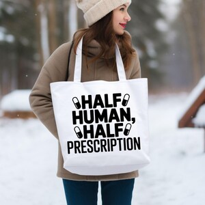 May include: White tote bag with the text "HALF HUMAN, HALF PRESCRIPTION" in black. The bag has two black pill graphics. The bag is carried by a person wearing a tan coat and a cream-colored beanie.