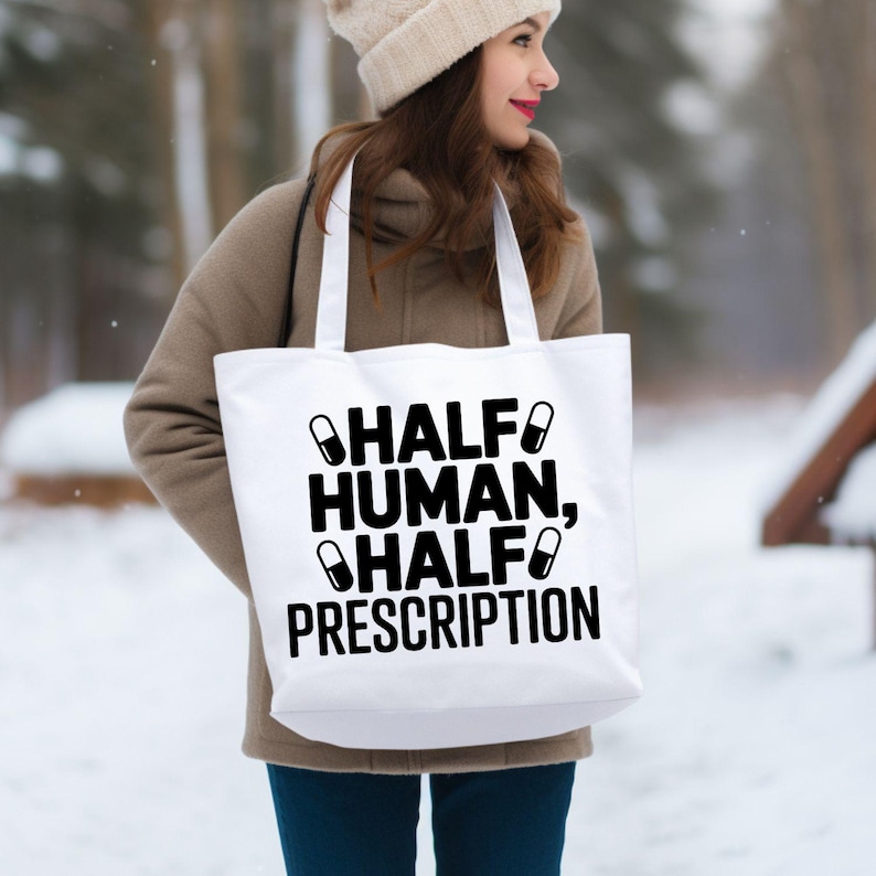 May include: White tote bag with the text "HALF HUMAN, HALF PRESCRIPTION" in black. The bag has two black pill graphics. The bag is carried by a person wearing a tan coat and a cream-colored beanie.