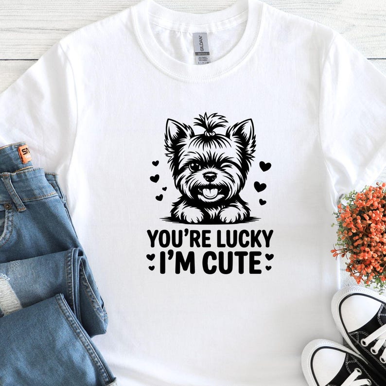 May include: White t-shirt featuring a black and white illustration of a winking Yorkshire Terrier with hearts. The text "YOU'RE LUCKY I'M CUTE" is printed below the dog. The shirt is paired with blue jeans and black and white sneakers.