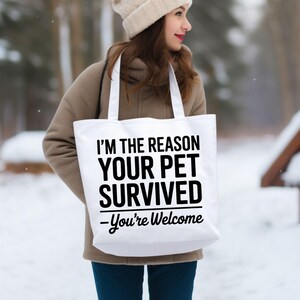 May include: A white tote bag with black text that reads "I'M THE REASON YOUR PET SURVIVED - You're Welcome." The bag has white handles and is being held by a person wearing a tan coat and hat. The background is a snowy outdoor scene.
