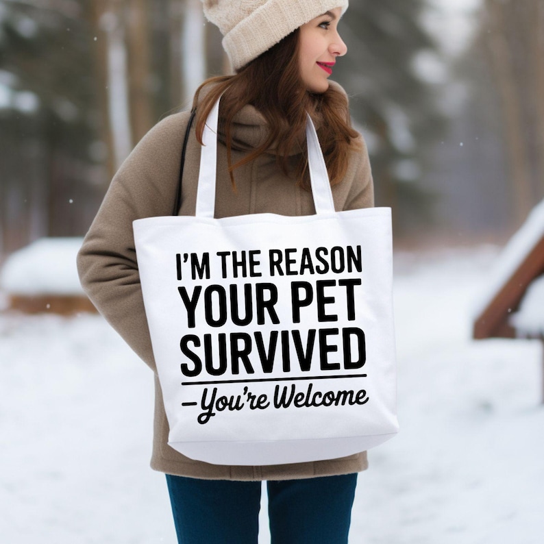 May include: A white tote bag with black text that reads "I'M THE REASON YOUR PET SURVIVED - You're Welcome." The bag has white handles and is being held by a person wearing a tan coat and hat. The background is a snowy outdoor scene.