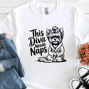 May include: White t-shirt with a black graphic of a Yorkshire Terrier wearing a robe, with the text "This Diva Needs Naps". The dog has a bow in its hair. The shirt is paired with blue jeans and black and white sneakers.