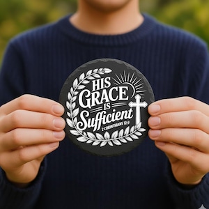 May include: A round, black slate coaster with white text that reads "HIS GRACE IS Sufficient 2 Corinthians 12:9". The design includes a cross, sun rays, and a laurel wreath. The coaster is held in front of a blurred green background.