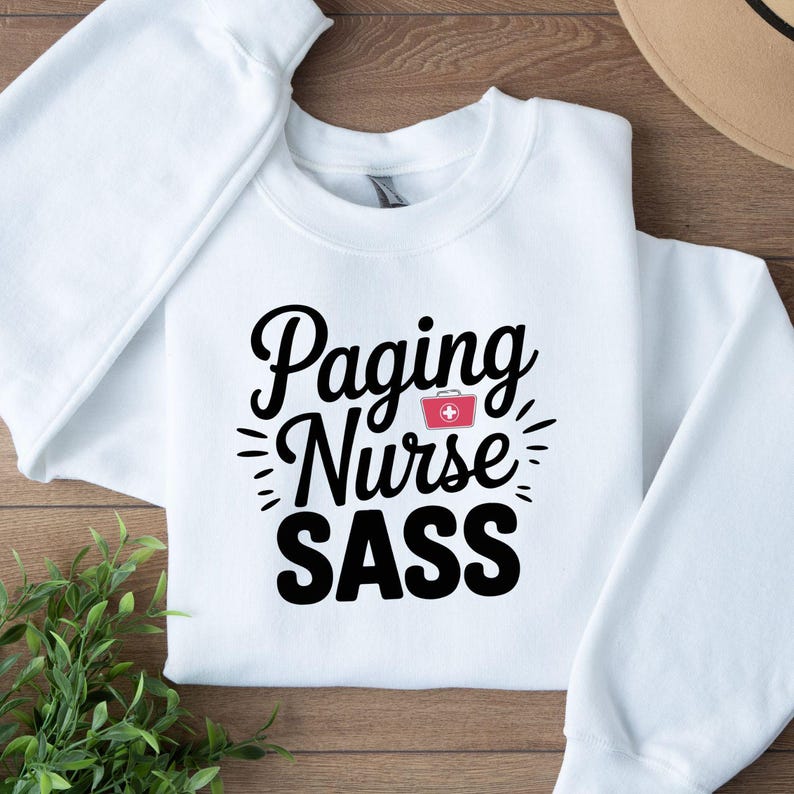 May include: White crewneck sweatshirt with the words "Paging Nurse SASS" in black script. The word "Nurse" is in a stylized font, and a small red first aid kit graphic is included. The sweatshirt is folded on a wooden surface.