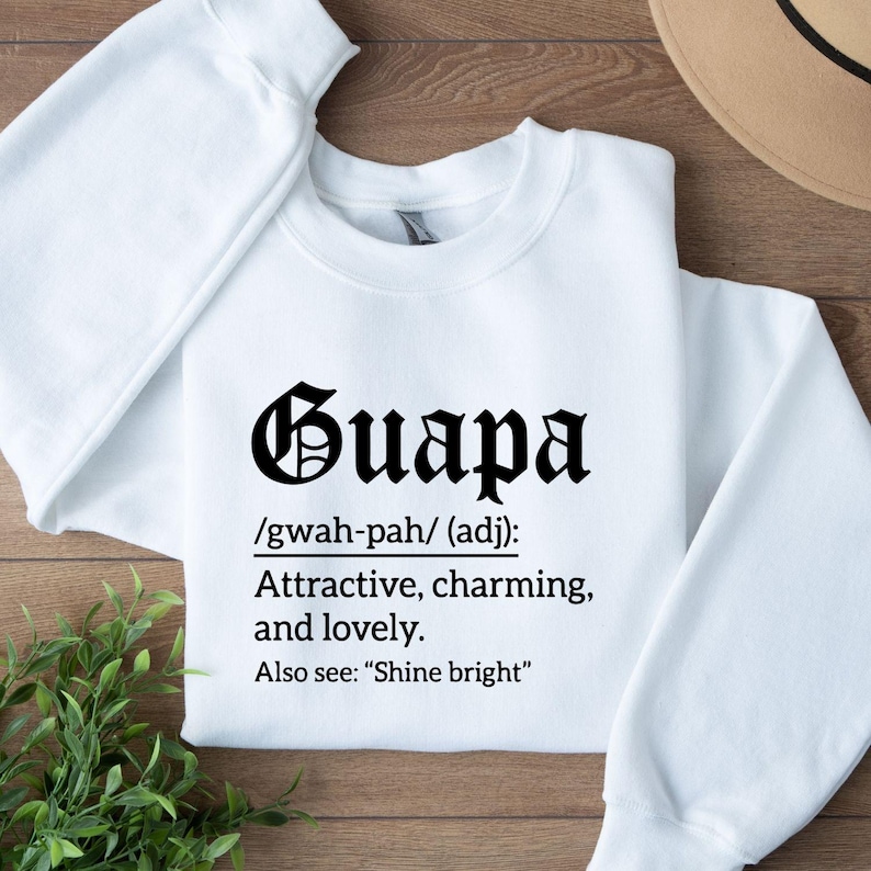 May include: A white sweatshirt with the word "Guapa" in a black, ornate font. Below the word is a definition: "Attractive, charming, and lovely." The sweatshirt also includes the pronunciation and a reference to "Shine bright."