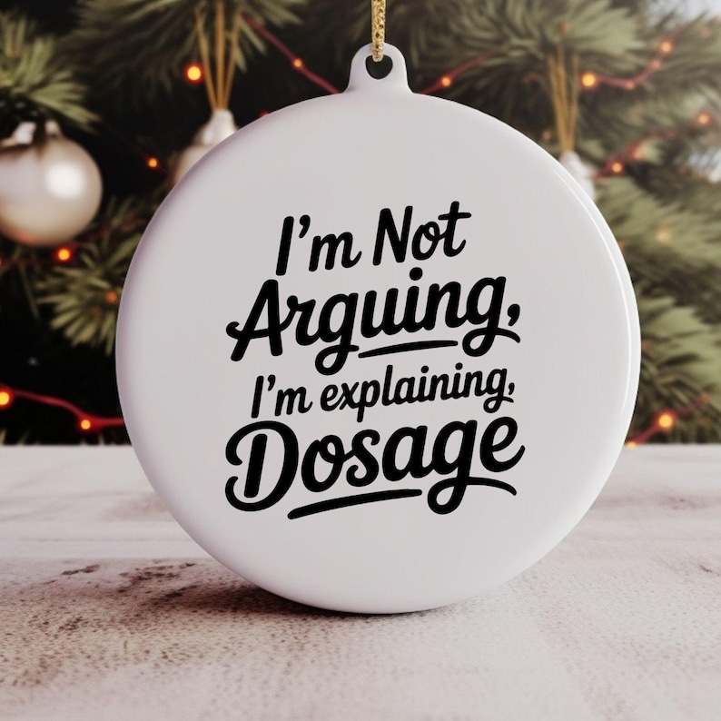May include: White round Christmas ornament with the text "I'm Not Arguing, I'm explaining, Dosage" in black script. The ornament hangs from a gold string, with a blurred Christmas tree in the background.