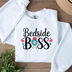 May include: White crewneck sweatshirt with the text "Bedside Boss" in black script. The design includes a cartoon character with a red hat and blue accents, along with red and teal hearts.