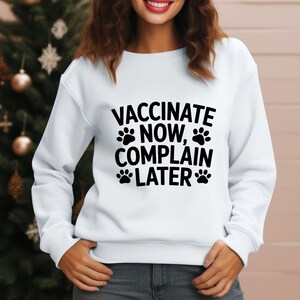 May include: White sweatshirt with the text "VACCINATE NOW, COMPLAIN LATER" in black, accompanied by paw prints. The sweatshirt is a crew neck style.