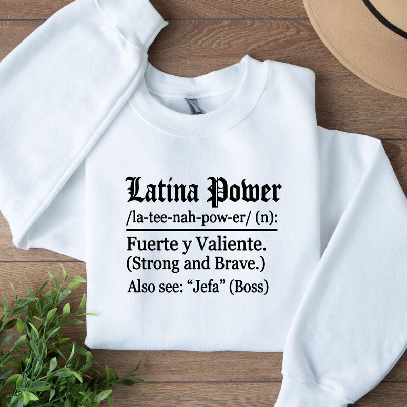 May include: White sweatshirt with the text "Latina Power" in a decorative font. The text includes a phonetic pronunciation and the Spanish translation "Fuerte y Valiente" (Strong and Brave).
