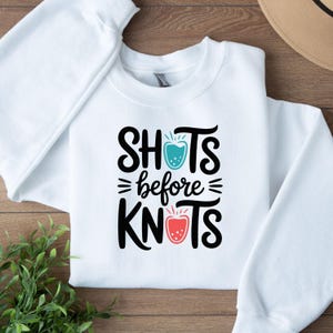 May include: White sweatshirt with the text "SHOTS before KNOTS" in black, with teal and red graphic accents. The sweatshirt is a crew neck style, with long sleeves. The design is centered on the front of the garment.