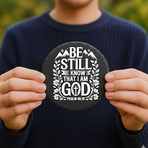 May include: A round, black slate coaster with white text that reads "BE STILL KNOW THAT I AM GOD" and "PSALM 46:10". The design includes mountain and floral elements. A decorative item for home or office.