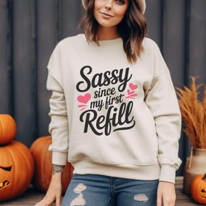 May include: A cream-colored sweatshirt with the text "Sassy since my first Refill" in black script and pink hearts. The sweatshirt is part of a fall-themed collection, with pumpkins in the background.