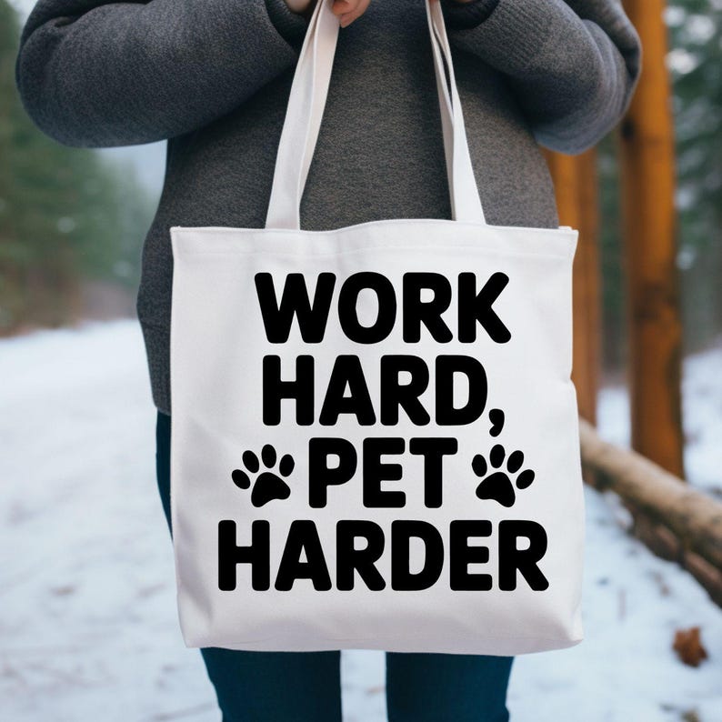May include: A white tote bag with black text that reads "WORK HARD, PET HARDER" and paw print graphics. The bag has two handles and is held by a person. The background is a blurred outdoor scene.