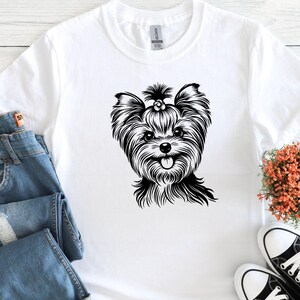 May include: White t-shirt featuring a black and white illustration of a Yorkshire Terrier dog. The dog has long, flowing fur, a flower in its hair, and a happy expression. The shirt is paired with blue jeans and black and white sneakers.