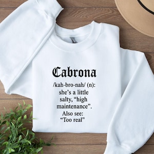 May include: White sweatshirt with the word "Cabrona" in a bold, black font. Below the word is a definition: "she's a little salty, 'high maintenance'". The sweatshirt also includes the phrase "Too real".