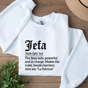 May include: White sweatshirt with the word "Jefa" in a bold, black font. Below the word is a definition: "The boss lady, powerful and in charge. Makes the rules, breaks barriers. Also see: 'La Patrona'"