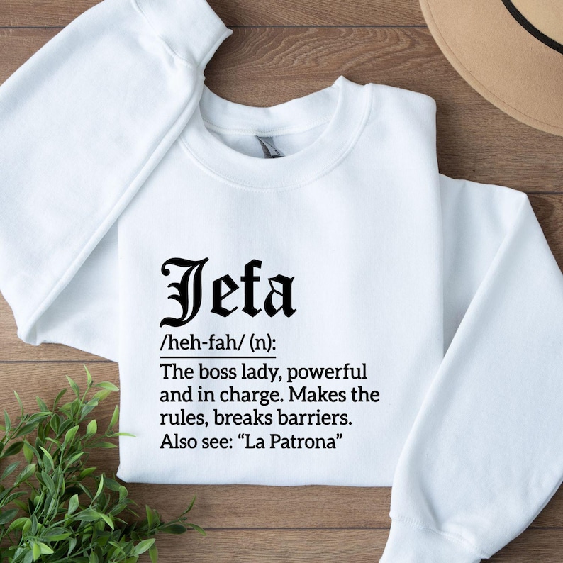 May include: White sweatshirt with the word "Jefa" in a bold, black font. Below the word is a definition: "The boss lady, powerful and in charge. Makes the rules, breaks barriers. Also see: 'La Patrona'"