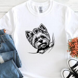 May include: White t-shirt featuring a black and white illustration of a Yorkshire Terrier dog peeking out of a pocket. The dog's fur is detailed, and the design is centered on the shirt. The shirt is paired with blue jeans and sneakers.