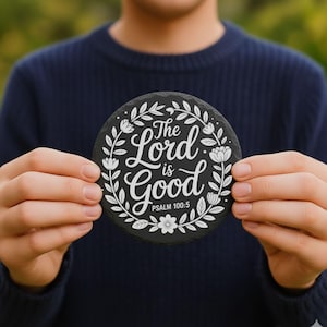 May include: A circular black coaster featuring white floral and leaf designs. The text "The Lord is Good" is written in a script font, with "Psalm 100:5" below. The coaster is held against a blurred green backdrop.