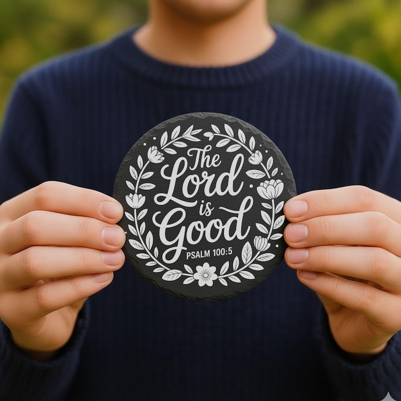 May include: A circular black coaster featuring white floral and leaf designs. The text "The Lord is Good" is written in a script font, with "Psalm 100:5" below. The coaster is held against a blurred green backdrop.