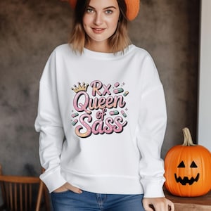 May include: White sweatshirt with the text "Rx Queen of Sass" in pink, yellow, and green. The design includes a crown and pill graphics. A carved orange pumpkin sits to the right of the sweatshirt.