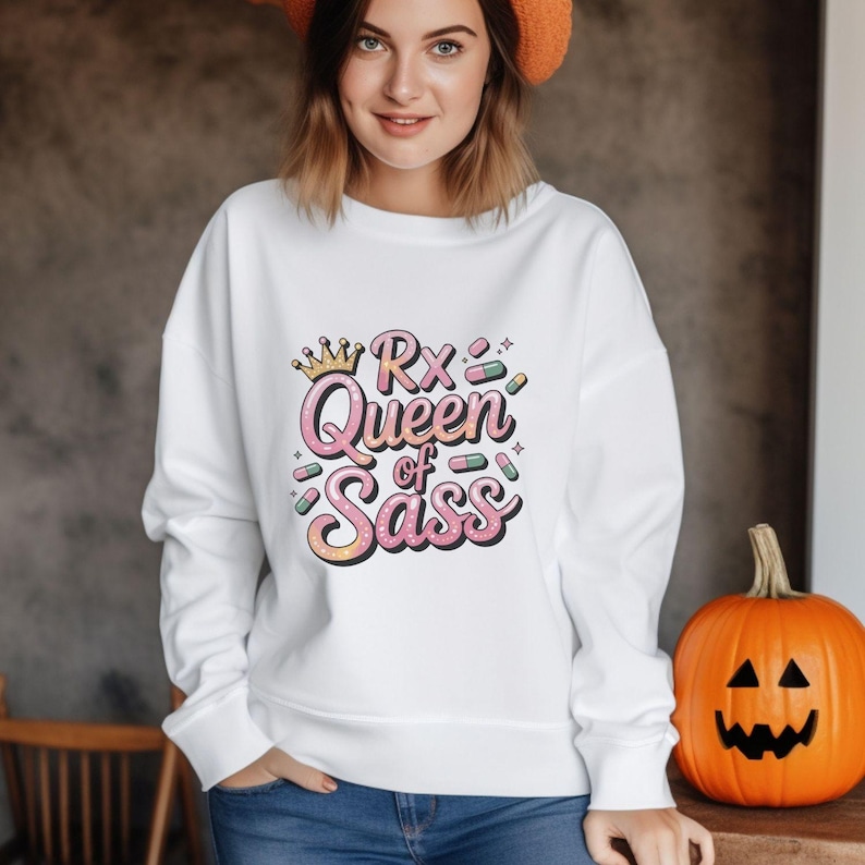 May include: White sweatshirt with the text "Rx Queen of Sass" in pink, yellow, and green. The design includes a crown and pill graphics. A carved orange pumpkin sits to the right of the sweatshirt.