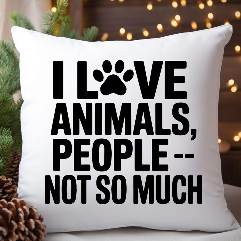 May include: White square throw pillow with the text "I LOVE ANIMALS, PEOPLE -- NOT SO MUCH" in bold black letters. A black paw print replaces the "O" in "LOVE". The pillow is on a white surface with a blurred background.