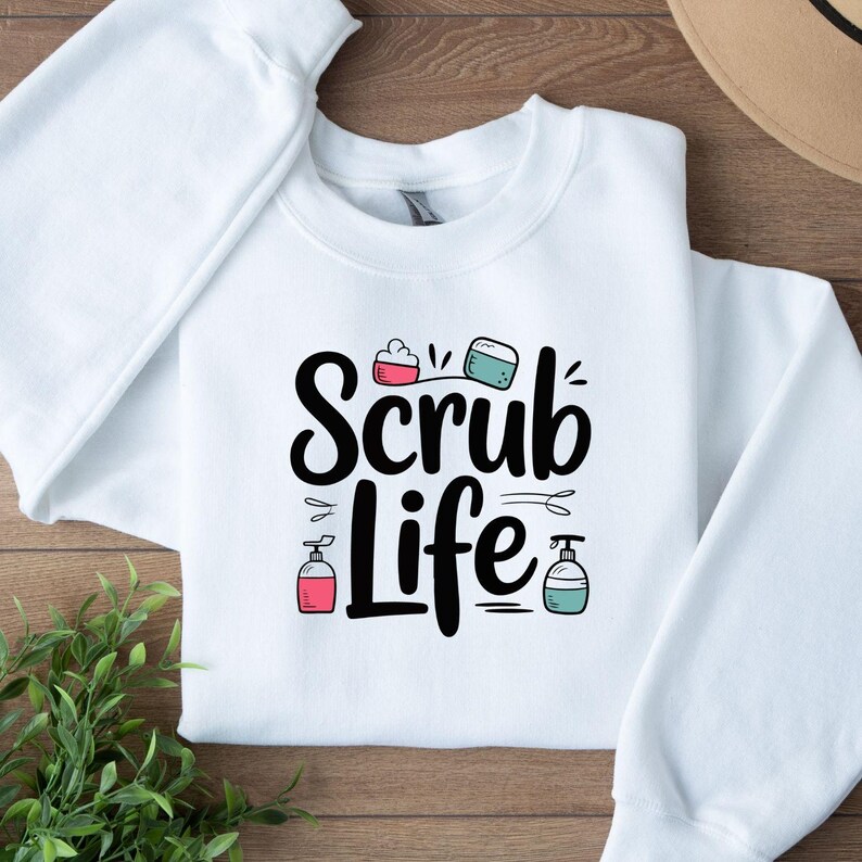 May include: White crewneck sweatshirt with the words "Scrub Life" in black script. The design includes illustrations of soap, soap dispensers, and other related items in pink and teal. The sweatshirt is folded on a wooden surface.