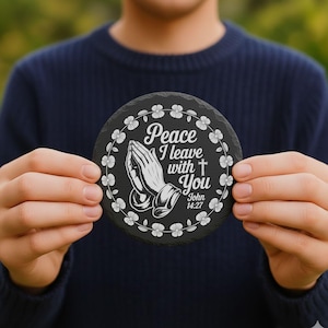 May include: Round black slate coaster with white floral border and text "Peace I leave with You" with praying hands and a cross. The text also includes "John 14:27".