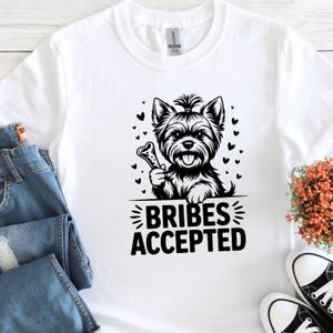 May include: White t-shirt featuring a black and white illustration of a Yorkshire Terrier holding a bone. The text "BRIBES ACCEPTED" is below the dog. The design includes small heart shapes.