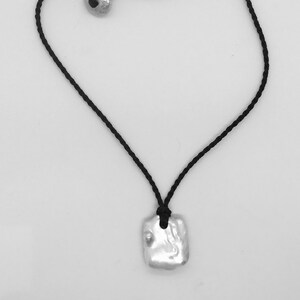 May include: A necklace featuring a black braided cord with a silver-toned, irregular rectangular pendant. The cord includes a matching silver-toned circular bead closure, creating a simple design that secures the pendant at the centre of the cord.