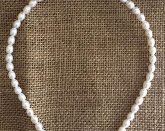 May include: A white pearl headband. The headband is made of small, oval-shaped pearls strung together. The pearls are a bright white colour and are arranged in a continuous line. The background is a textured, brown fabric.