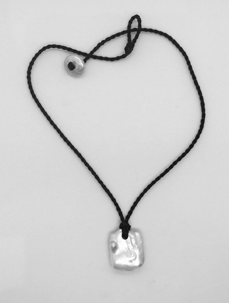 May include: A necklace featuring a black braided cord with a silver-toned, irregular rectangular pendant. The cord includes a matching silver-toned circular bead closure, creating a simple design that secures the pendant at the centre of the cord.