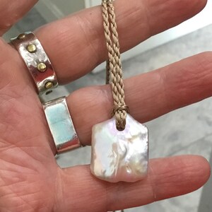 May include: A pendant featuring an irregular, iridescent white baroque pearl suspended from a braided tan cord. The pearl has a flat, rectangular shape with a soft lustre. Two silver rings are also visible, one with gold-toned studs.